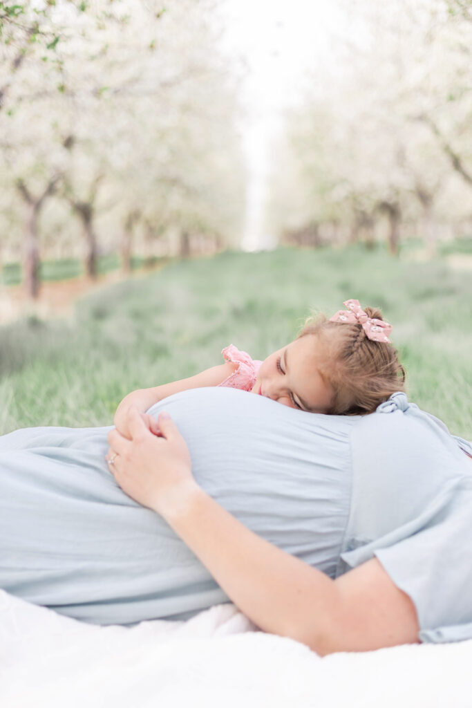 A child poses with her pregnant mother's baby bump in a Utah orchard flowering with white blossoms in a spring family and maternity photo session by a Utah County photographer