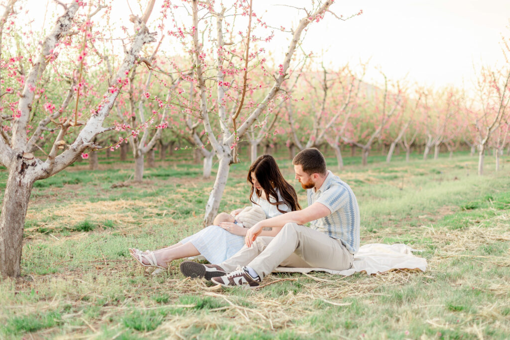 Parents sitting in blooming spring orchard holding their newborn baby in Utah County, soft natural light family photography in Provo Utah blossom trees