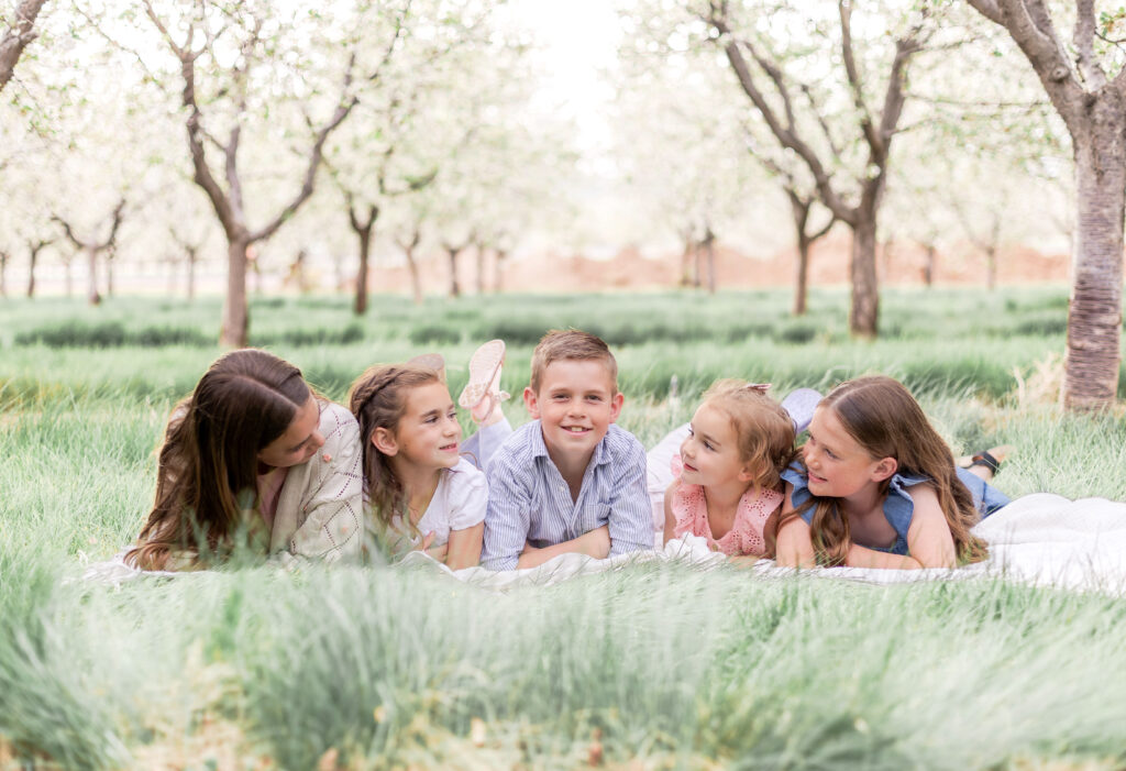 Young siblings laughing together while laying among blossoming trees in a Utah orchard during a Utah County family photo session.