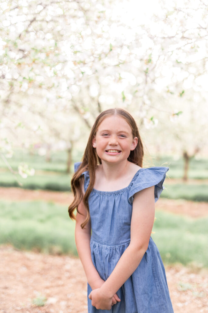 Portrait of a little girl standing in a sunlit Utah orchard with soft greenery and timeless white blossoms in the background.