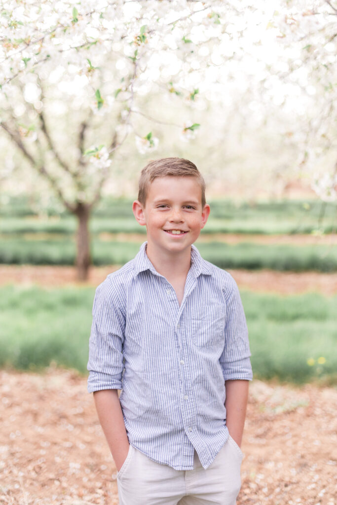 Young boy smiling during a portrait session in a Utah orchard with timeless blossoming trees behind him.