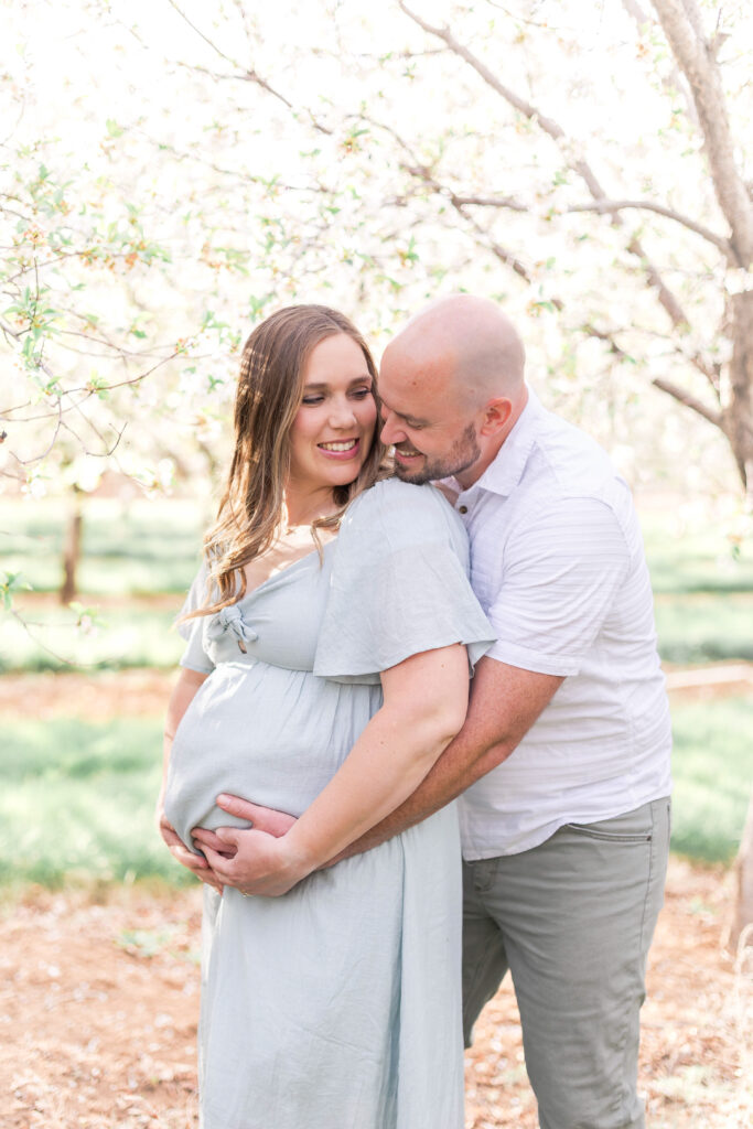 Expecting couple laughing together while standing in a Utah orchard during a maternity session in Utah County.
