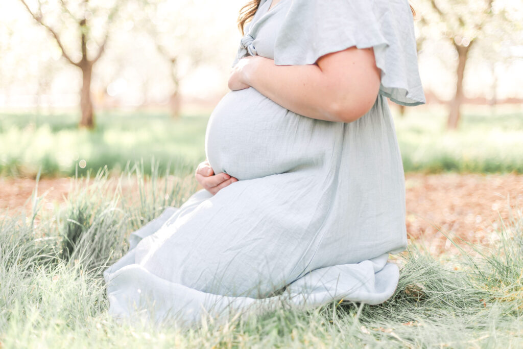 Pregnant woman photographed in a Utah orchard with trees and soft natural light during a maternity session.