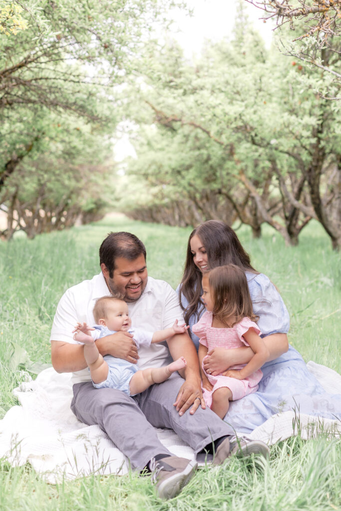 A candid moment of a family smiling at each other in a Utah orchard, with soft spring light glowing through the trees behind them.