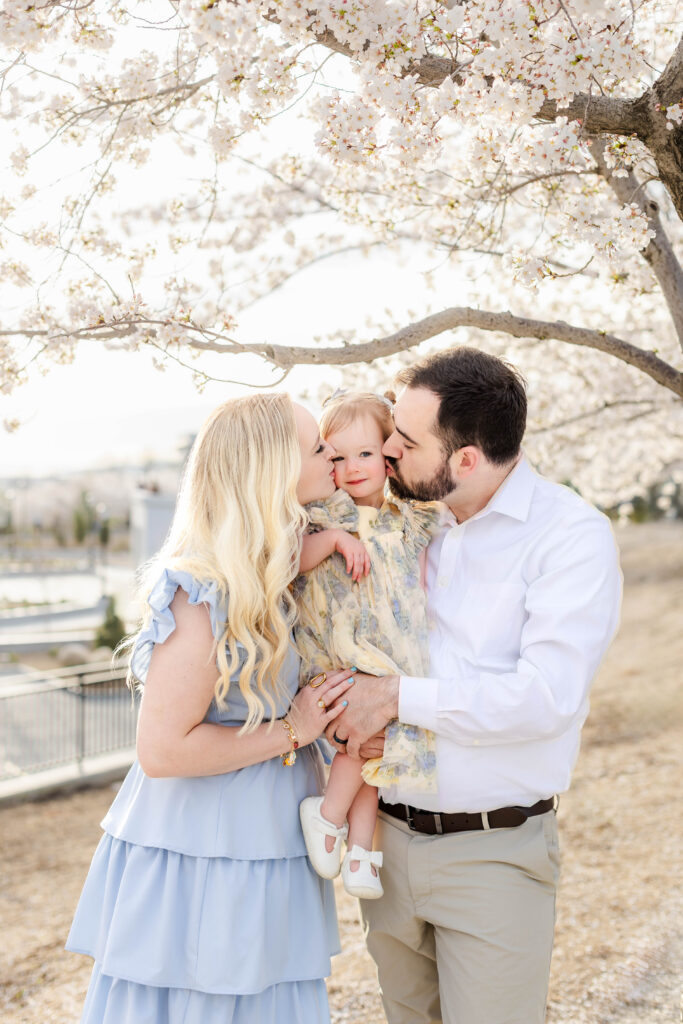 Utah cherry blossom family photos of parents kissing their young daughter under blooming trees in soft spring light
