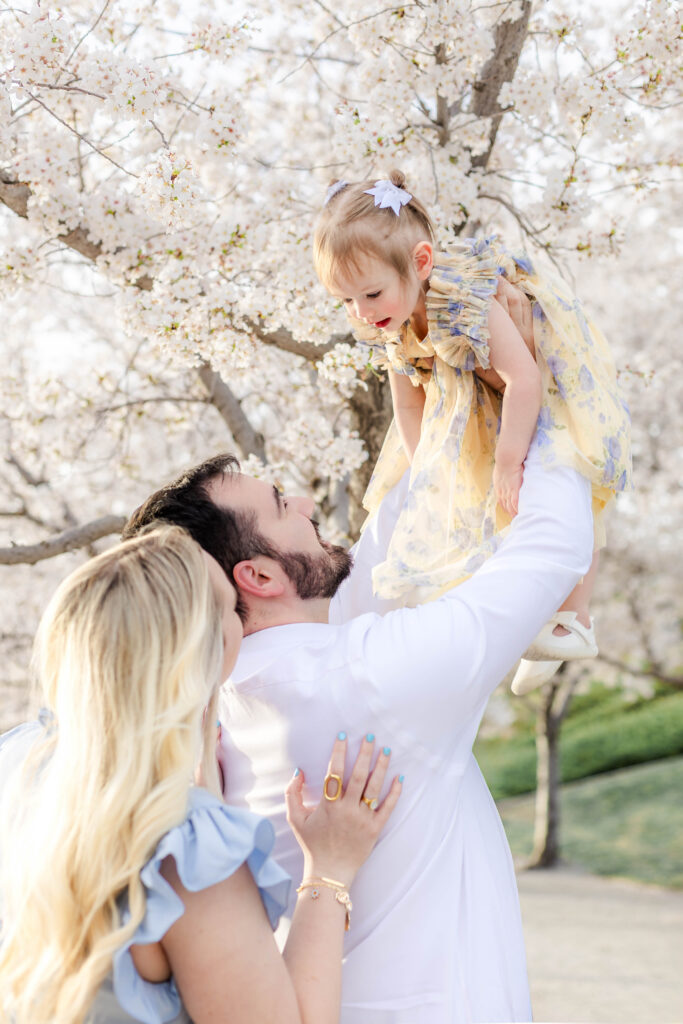 A dad lifts toddler up in the air while mom stand behind among blooming cherry blossoms in Utah