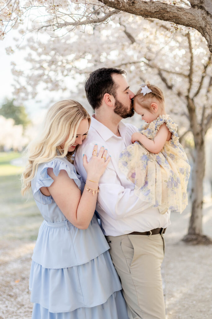 Mom hugs dad from behind while dad kisses baby girl for a family photo session under the cherry blossoms in Utah