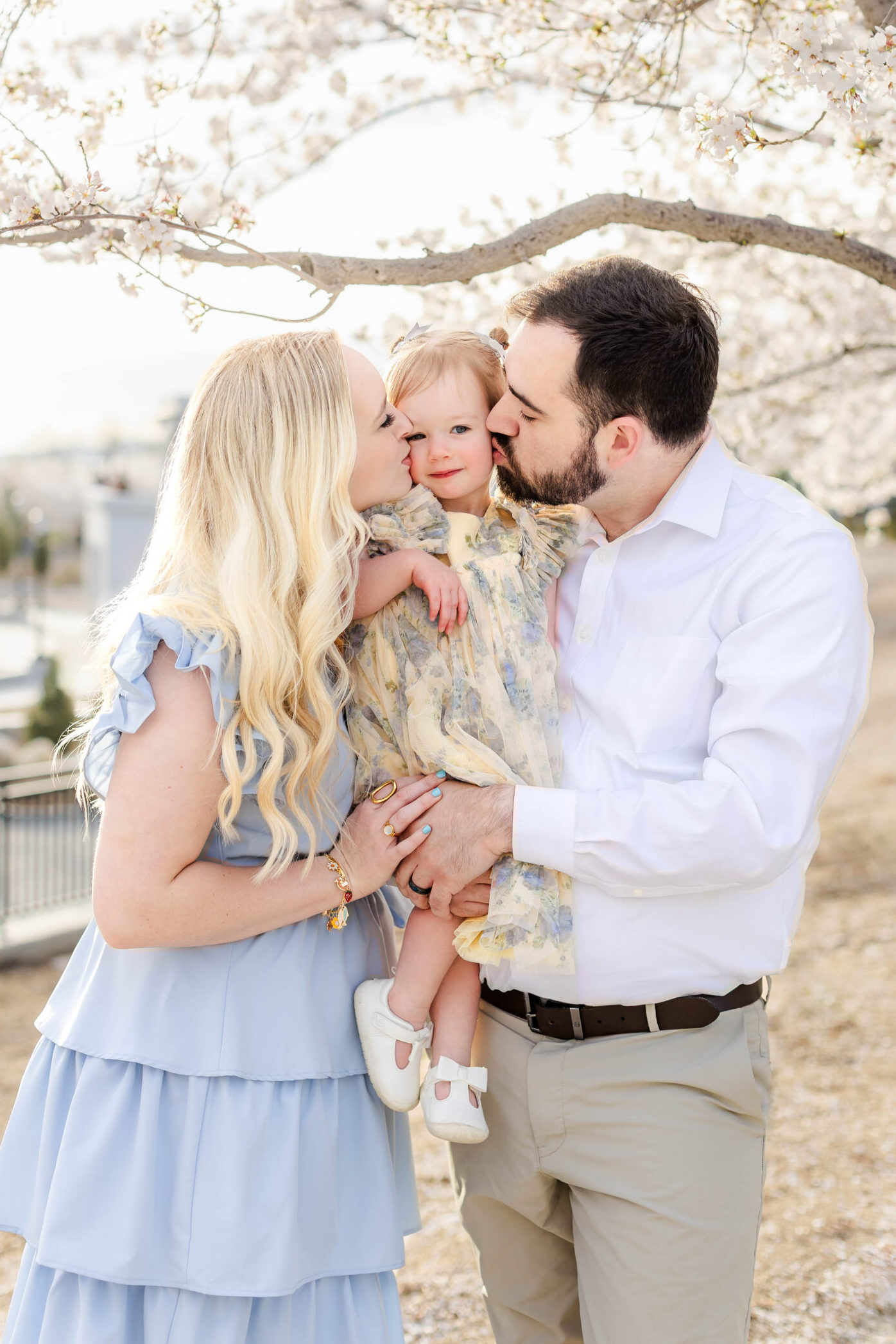 Utah cherry blossom family photos of parents kissing their young daughter under blooming trees in soft spring light